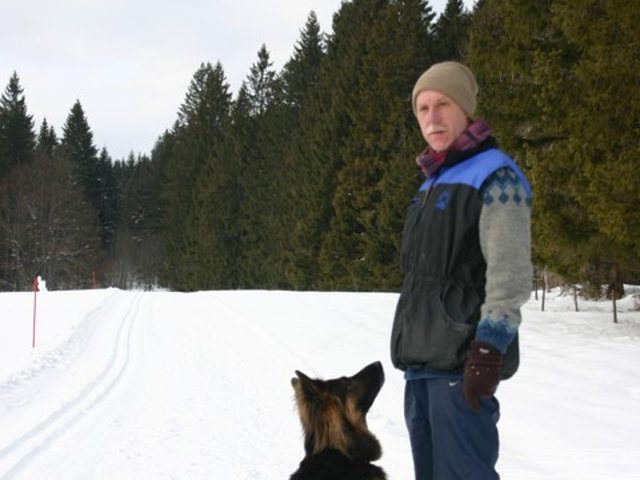 Mann und Schäferhund im Schnee vor einem Nadelwald