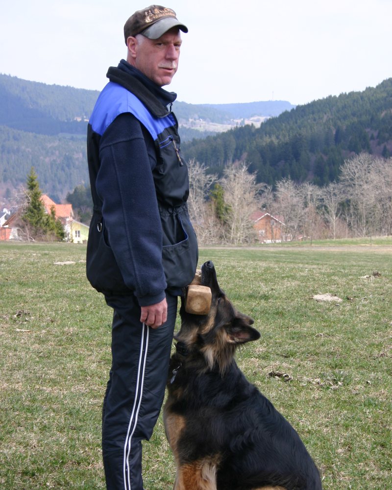 Mann steht auf einer Wiese, neben ihm sitzt ein Schäferhund
